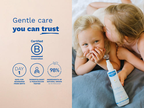 Two young children lying on a bed, one whispering to the other, with a Mustela spray bottle in the foreground and blue text on the left side. - Mustela USA - 5