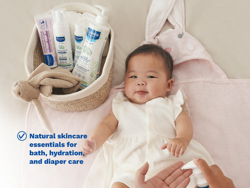 Mustela USA | Smiling baby lying on a pink blanket next to a basket of Mustela skincare products including Gentle Cleansing Gel and Diaper Rash Cream, with a parent applying lotion. - Mustela USA - 2